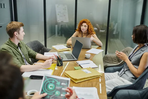 Рыжая деловая женщина разговаривает с коллегами возле документов и ноутбуков в офисе, обсуждая стартап — стоковое фото