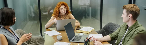 Redhead team lead talking to managers near documents and laptops in office meeting room, banner — Stock Photo
