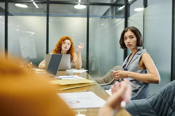 Young businesswomen looking at blurred colleague near laptops and documents in meeting room — Stock Photo