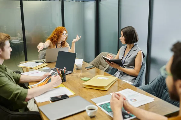Redhead businesswoman pointing with finger near colleagues, meeting room, laptops, documents, graphs — Stock Photo