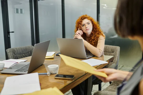 Thoughtful redhead entrepreneur near blurred manager with envelopes, office, laptops, documents — Stock Photo