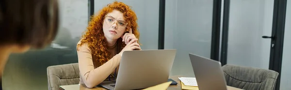 Серьезный рыжий предприниматель смотрит на размытого бизнес-партнера рядом с ноутбуками в офисе, баннер — стоковое фото