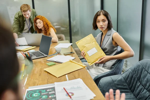 Young businesswoman with envelope near colleagues talking in meeting room, laptops, graphs — Stock Photo
