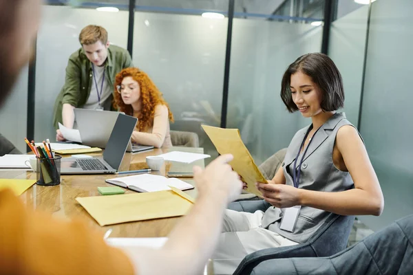 Mulher feliz com envelope perto de colegas que trabalham na inicialização, sala de reuniões, laptops, documentos — Fotografia de Stock