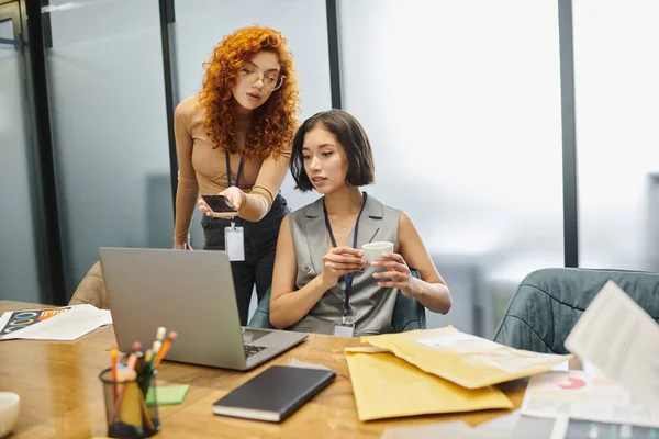 Redhead businesswoman with smartphone near colleague with coffee cup working on laptop in office — Stock Photo