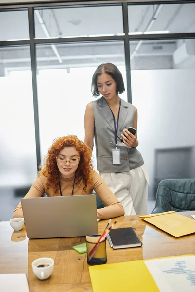 Young team lead with smartphone near redhead manager working on laptop in modern office — Stock Photo