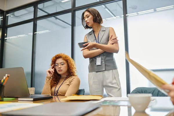 Redhead thoughtful businesswoman looking at laptop near team lead with smartphone in modern office — Stock Photo