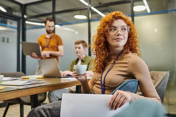 Redhead team lead with coffee cup looking away, businessmen talking on blurred background in office — Stock Photo