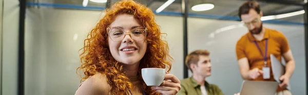 Portrait d'entrepreneur roux dans des lunettes, avec tasse de café près de collègues flous, bannière — Photo de stock