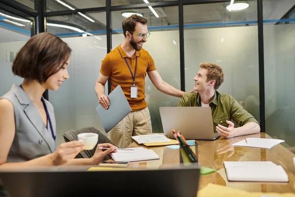 Cheerful bearded team lead with laptop touching shoulder of colleague working in meeting room — Stock Photo