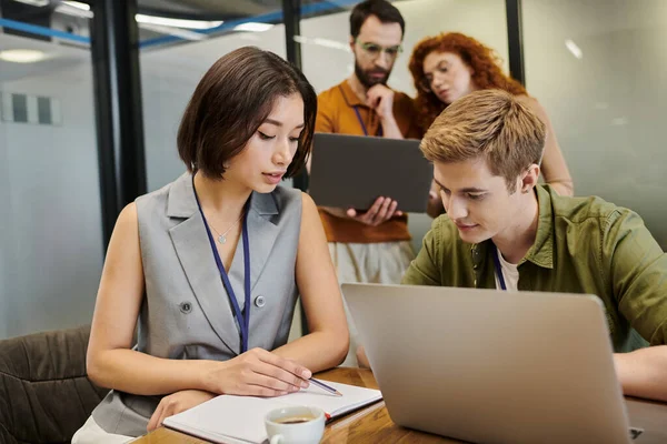 Joven empresaria apuntando a cuaderno cerca de colega y portátil, trabajo en equipo, proyecto de inicio — Stock Photo