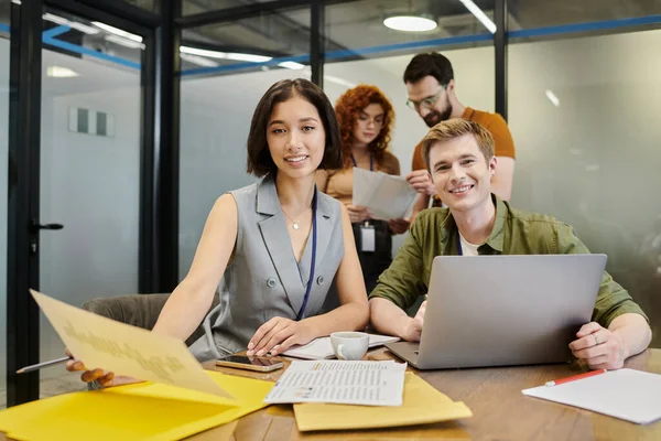 Portrait of young entrepreneurs looking at camera near laptop, blurred colleagues talking in office — Stock Photo
