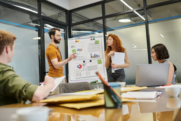 Bearded entrepreneur pointing near infographics on flip chart near colleagues in meeting room — Stock Photo