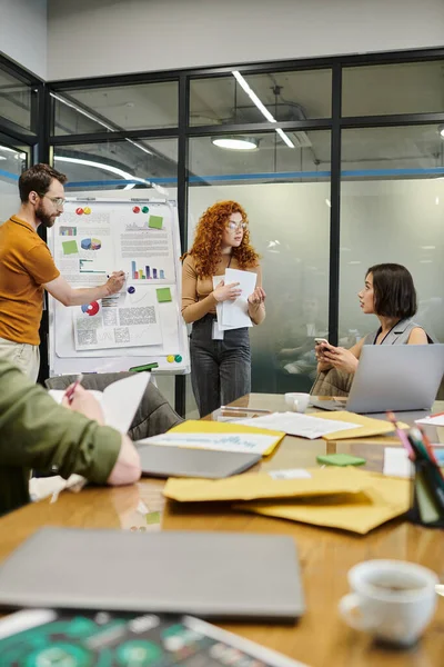 Uomo d'affari barbuto indicando grafici su flip chart e team creativo discutendo di start-up in ufficio — Foto stock