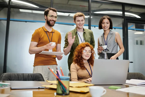 Счастливый бизнесмен машет рукой во время видеозвонка на ноутбук рядом с коллегами в современном офисе — стоковое фото