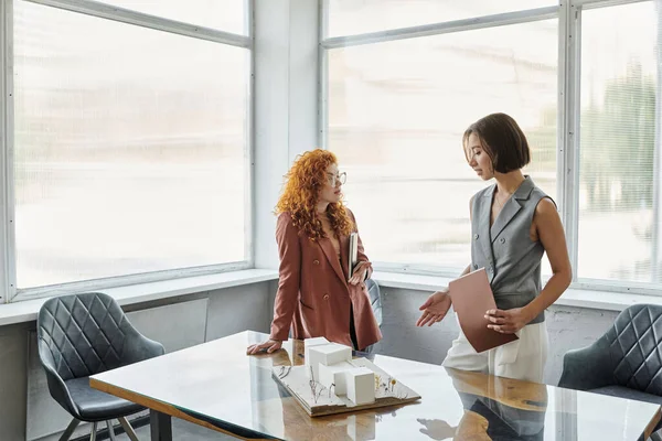 Giovane donna d'affari che parla con redhead designer vicino al modello architettonico in ufficio moderno — Foto stock