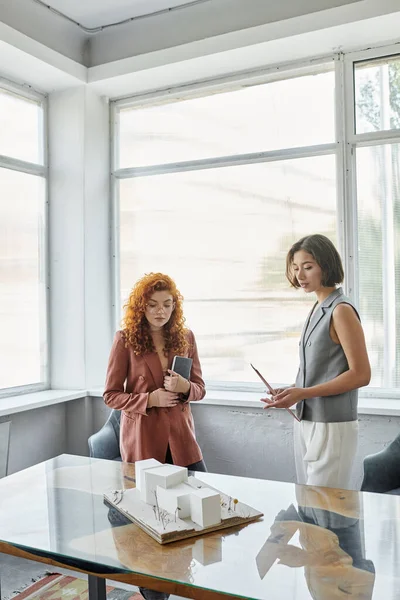 Redhead architect and young investor talking near building model in office, project presentation — Stock Photo