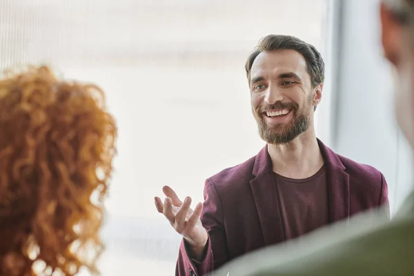 Улыбающийся бородатый бизнесмен разговаривает и жестикулирует рядом с рыжей женщиной на размытом переднем плане в офисе — стоковое фото