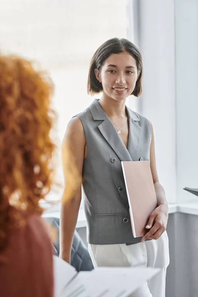 Jeune femme d'affaires avec dossier souriant près d'un collègue rousse au premier plan flou au bureau — Photo de stock