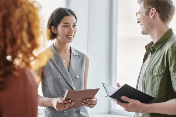 Young entrepreneur with folder smiling near business partner with notebook in contemporary office — Stock Photo