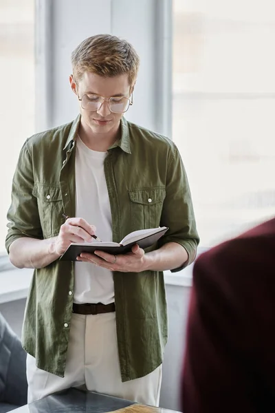 Serious businessman in eyeglasses writing in notebook on blurred foreground in modern office — Stock Photo