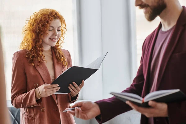 Positive redhead businesswoman looking at documents near colleague in coworking office — Stock Photo