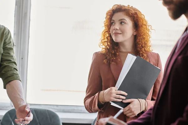 Young redhead businesswoman with documents smiling near colleagues in contemporary office — Stock Photo