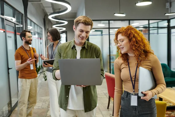 Redhead businesswoman pointing at laptop near colleague, coworkers talking on background in office — Stock Photo
