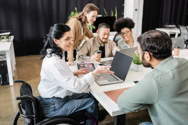 Cheerful team lead in wheelchair pointing at laptop, sharing ideas with man near business people — Stock Photo