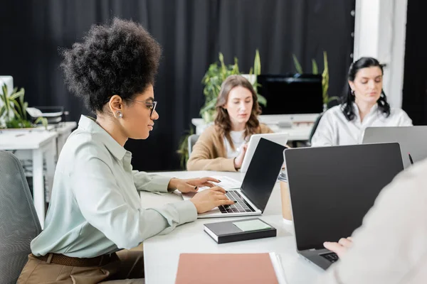 Vue latérale d'une femme d'affaires afro-américaine utilisant un ordinateur portable près de collègues féminines au bureau — Photo de stock