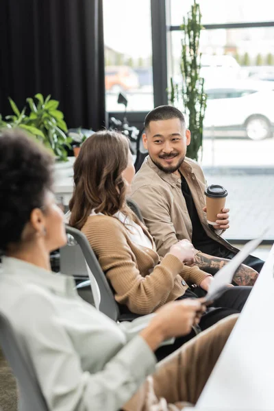 Joyeux asiatique homme d'affaires tenant café pour aller et bavarder avec une collègue féminine dans le coworking moderne — Photo de stock