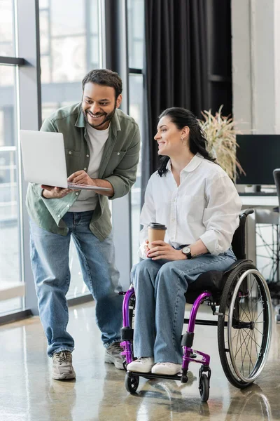 Cheerful indian businessman showing startup project on laptop to disabled woman in wheelchair — Stock Photo