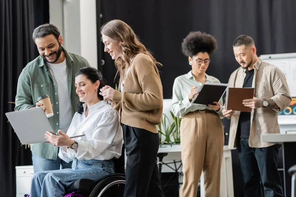 Disabled woman in wheelchair discussing startup project on laptop with happy interracial colleagues — Stock Photo