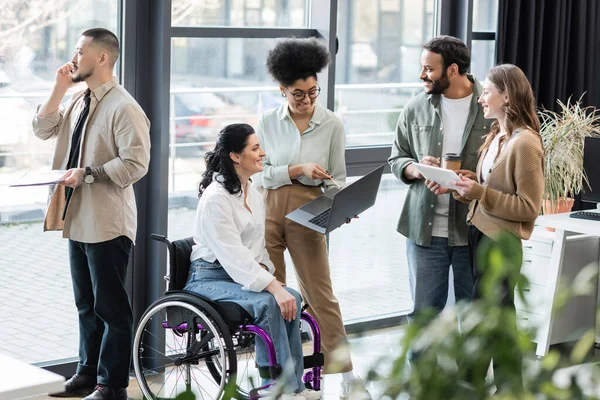 Cheerful interracial business people and disabled woman in wheelchair using devices in coworking — Stock Photo