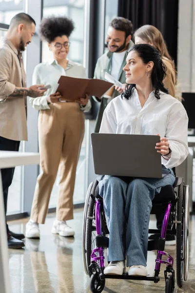 Femme d'affaires handicapée en fauteuil roulant utilisant un ordinateur portable près d'une équipe multiethnique floue, concept d'inclusion — Photo de stock