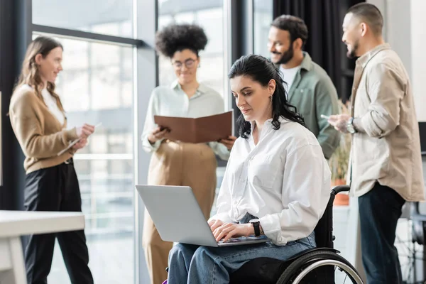 Disabled businesswoman in wheelchair using laptop near blurred multiethnic colleagues, inclusion — Stock Photo