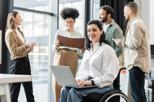 Mulher de negócios deficiente feliz em cadeira de rodas usando laptop perto de colegas multiétnicos borrados — Fotografia de Stock