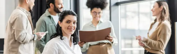 Happy businesswoman looking at camera near blurred multiethnic startup team in coworking, banner — Stock Photo