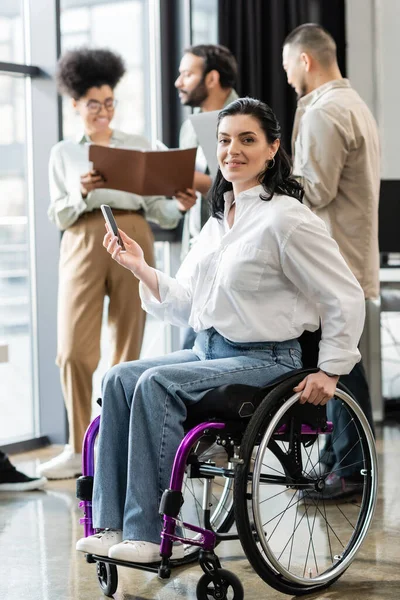 Felice donna d'affari disabile in sedia a rotelle guardando la fotocamera vicino a colleghi multietnici offuscati — Foto stock