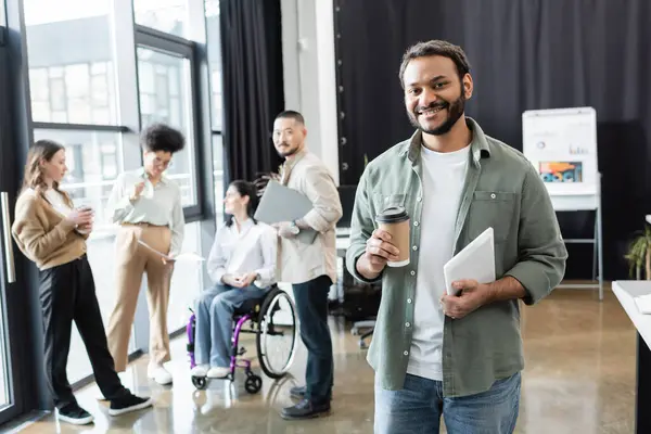 Cheerful indian office worker holding coffee to go and tablet near diverse startup team in coworking — Stock Photo