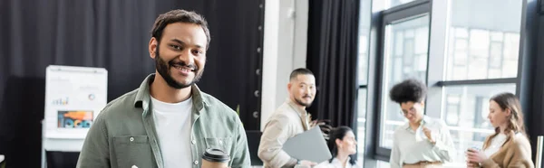 Щасливий індійський бізнесмен тримає каву і планшет біля різноманітної команди стартапів, банер — стокове фото