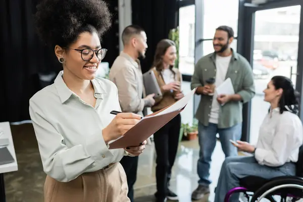 Cheerful african american woman taking notes and standing near interracial team in coworking — Stock Photo