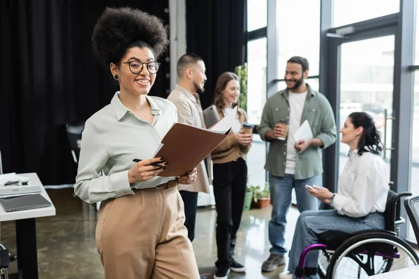 Cheerful african american woman holding folder and standing near interracial team in coworking — Stock Photo