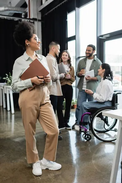 Pleased african american woman holding folder and standing near interracial team in coworking — Stock Photo