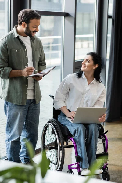 Happy indian man chatting with disabled businesswoman on wheelchair using laptop near window — Stock Photo