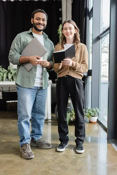 Cheerful woman with notebook and indian man with laptop looking at camera in office, coworking space — Stock Photo