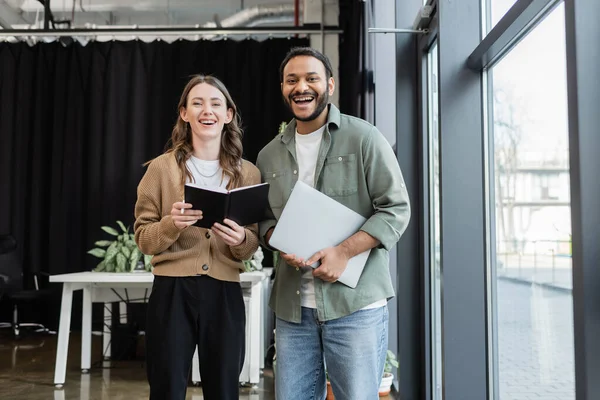 Joyful woman with notebook and indian man with laptop looking at camera in office, coworking space — Stock Photo