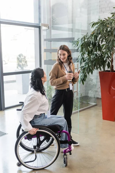 Conceito de inclusão, mulher feliz olhando para colega com deficiência em cadeira de rodas perto da porta de vidro — Fotografia de Stock