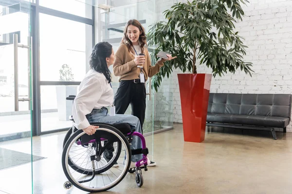 Inklusionskonzept, glückliche Frau zeigt weg und blickt behinderte Kollegin im Rollstuhl an — Stockfoto