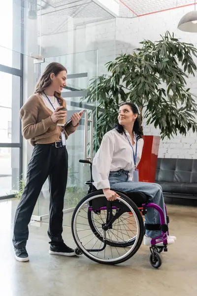 Concept d'inclusion, femme heureuse tenant un café et parlant avec un collègue handicapé en fauteuil roulant — Photo de stock